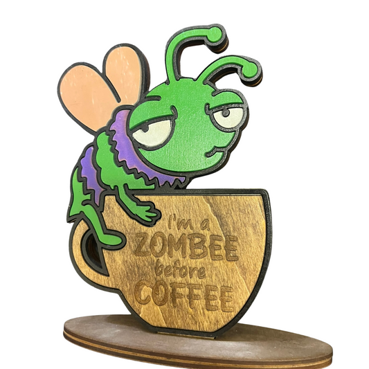 Wooden figurine of a green insect character on a coffee cup with 'I'm a zombee before coffee' text.